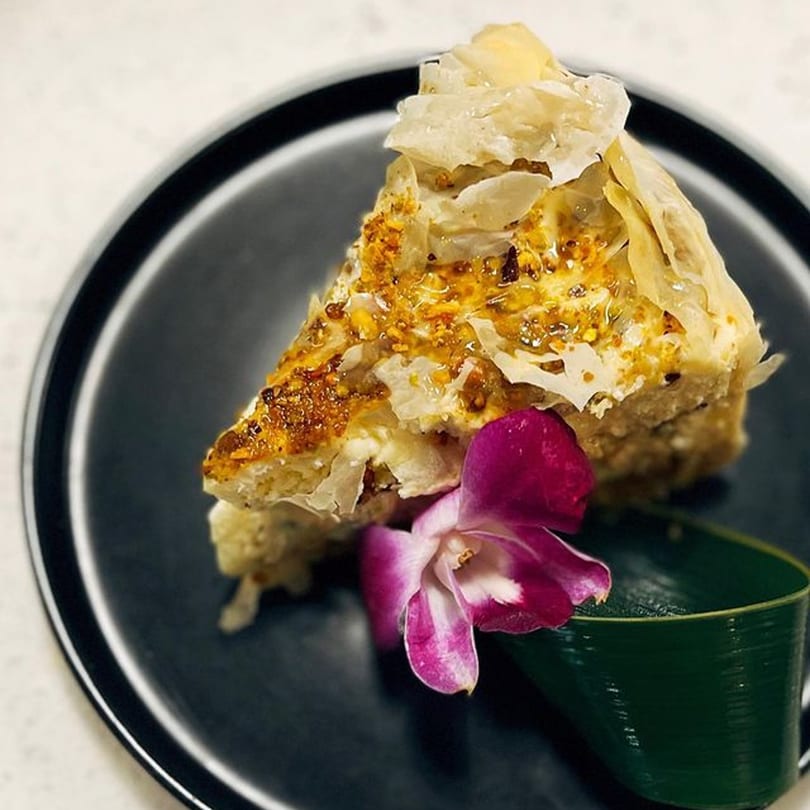 A slice of Japanese dessert with flaky pastry and a purple orchid on a black plate.